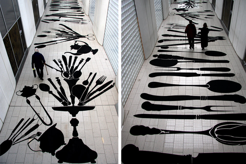 wo perspectives of a public walkway installation featuring large black cutlery and kitchen utensil silhouettes applied to the floor tiles. The designs, including forks, knives, teapots, and scissors, are scaled up dramatically, creating a striking visual effect as pedestrians walk over them.